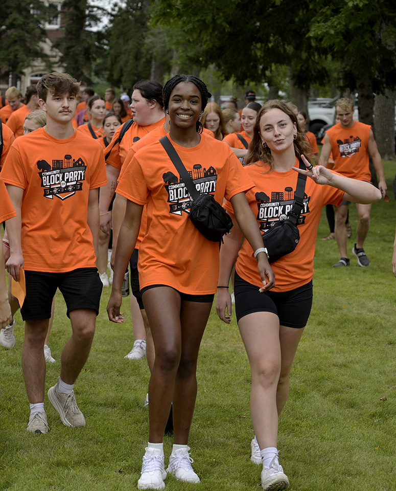 Two women in orange T-shirst that read "Community Block Party 2024" walk with other students in a crowd. 