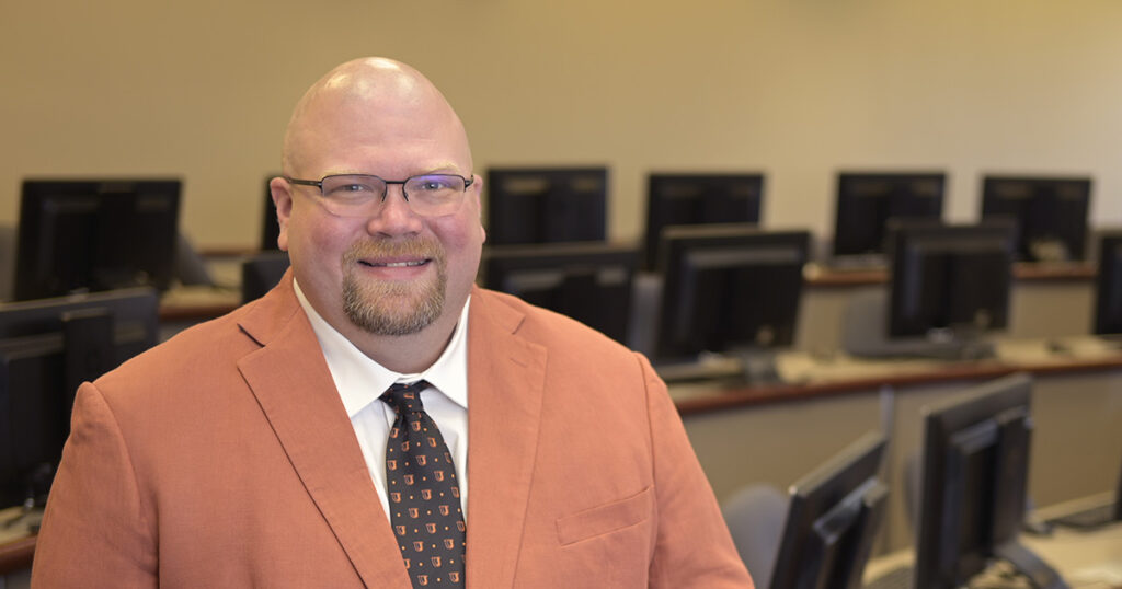 Dr. Jakob Barnard wears an apricot suit in this portrait while standing in front of a row of workstations in a computer lab. 