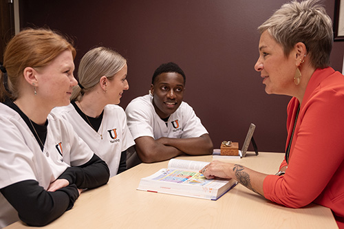 Three students in white nursing scrubs listen to a woman speaking from the other side of a desk. 