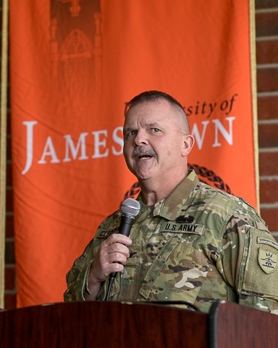 A man in a US Army National Guard camouflage uniform speaks into a microphone in front of an orange flag that says "University of Jamestown" in white letters