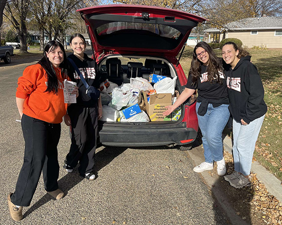 A group of four women stand around the open liftgate of a red car and show the bags of donated food they collected for a food drive. 