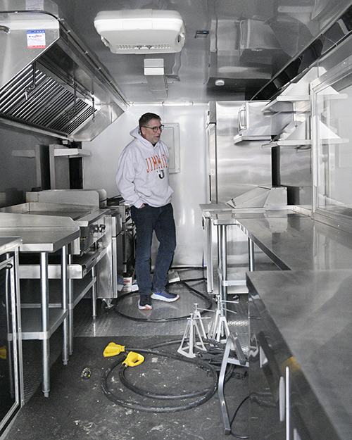 A man in a white hooded sweatshirt and jeans looks around a kitchen inside a trailer. All surfaces are stainless steel and he is facing a refrigerator. 