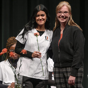 A woman in a white nursing scrub top holds a flower. She is next to another woman and they are both smiling for a photo. 