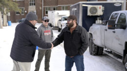 Two men in warm winter clothes shake hands in front of another man while a third man stands just past them. There is a truck towing a trailer, and the truck has 