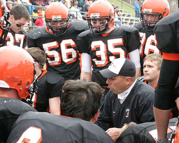 A group of football players wearing black jerseys and orange helmets stands around a man who is kneeling on the ground and coaching them. 